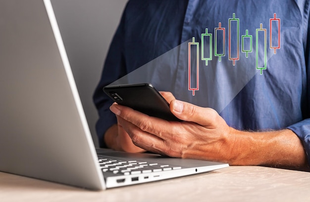 Hombre mirando el gráfico de velas en el teléfono mientras trabaja en la computadora portátil Análisis del mercado de valores Concepto de comercio de inversión financiera Empresario analizando el movimiento de precios