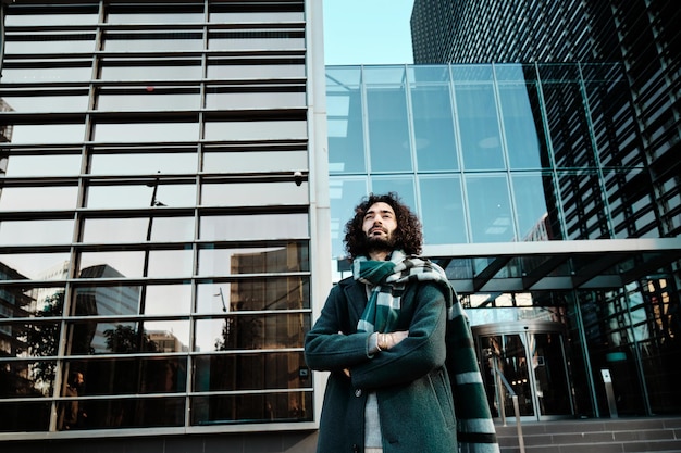 Foto hombre mirando hacia otro lado mientras posa con los brazos cruzados contra un edificio de oficinas en el distrito financiero.