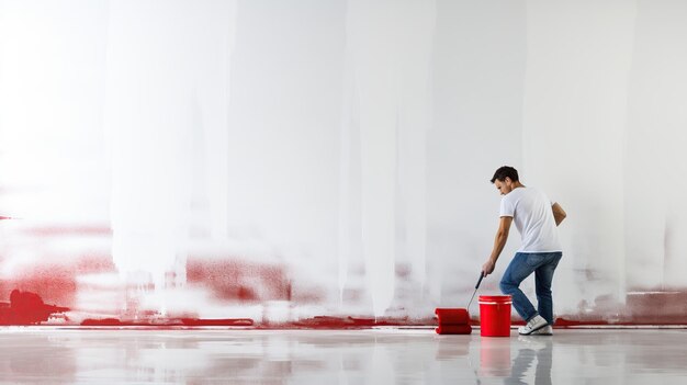 Un hombre mirando una pared en blanco con un rodillo de pintura y un balde aislado en una habitación blanca