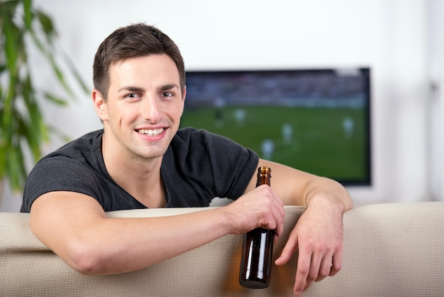 Hombre mirando un partido de fútbol en el sofá con una cerveza.