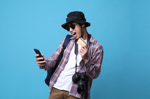 Foto hombre mochilero feliz sosteniendo un teléfono inteligente y una tarjeta de crédito sobre fondo de estudio azul.