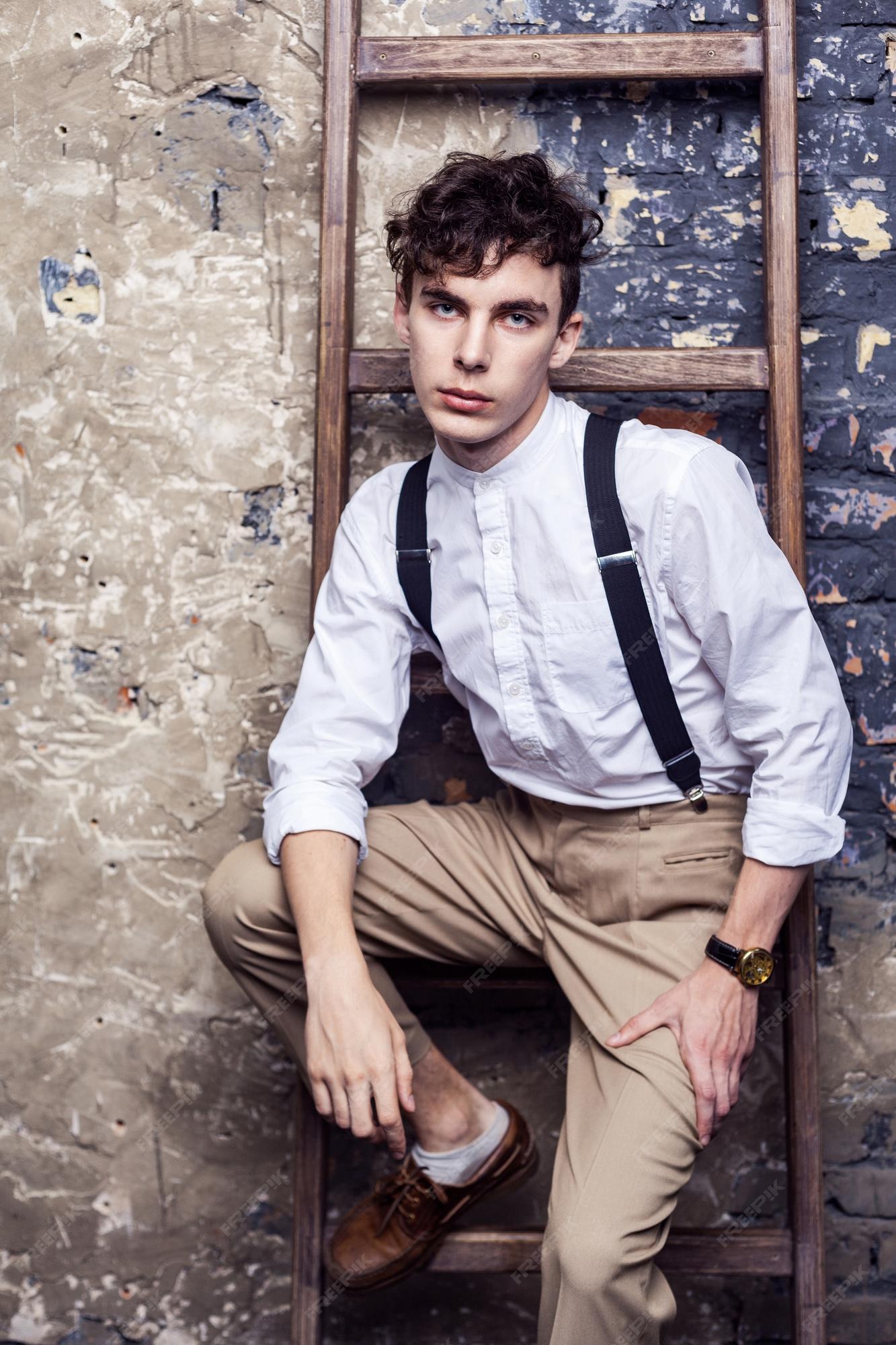 Hombre de moda en camisa blanca pantalón beige con tirantes sentado posando en la escalera de madera sobre de pared de ladrillo y mirando a cámara con cara seria.