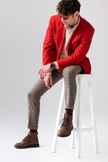 Hombre de moda con chaqueta roja y gafas de sol sentado en un taburete con la cabeza inclinada en gris