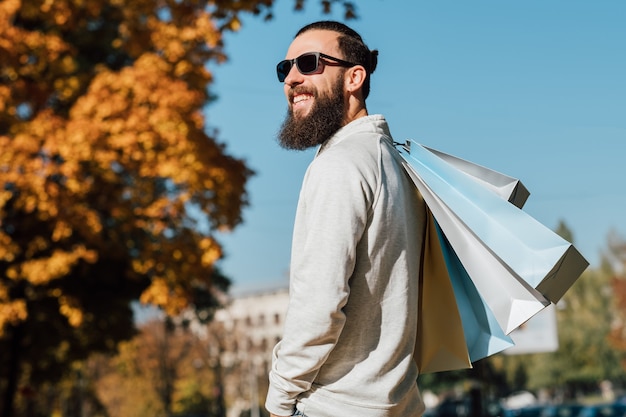 Hombre de moda estilo hipster barbudo con bolsas de compras disfrutando de día soleado de otoño