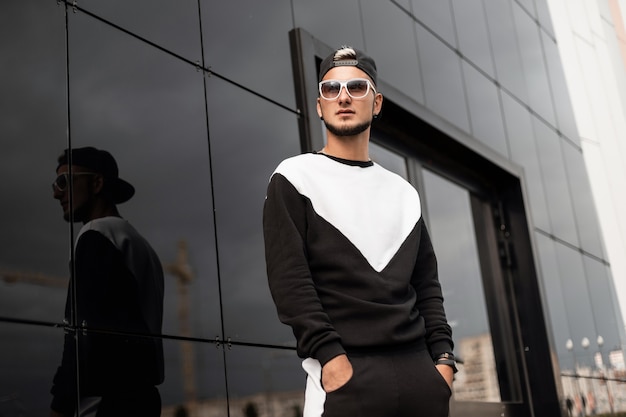 Hombre de moda con gafas de sol y gorra en sudadera camina en la ciudad