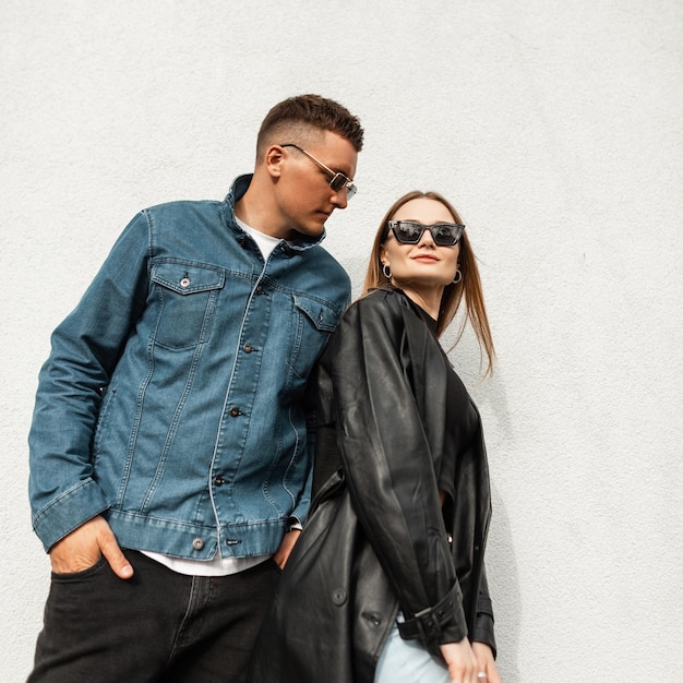 Foto hombre de moda y hermosa mujer con estilo con gafas de sol en ropa urbana de moda con chaqueta de jeans y soportes de abrigo de cuero y posa cerca de una pared blanca en la calle pareja de belleza de moda