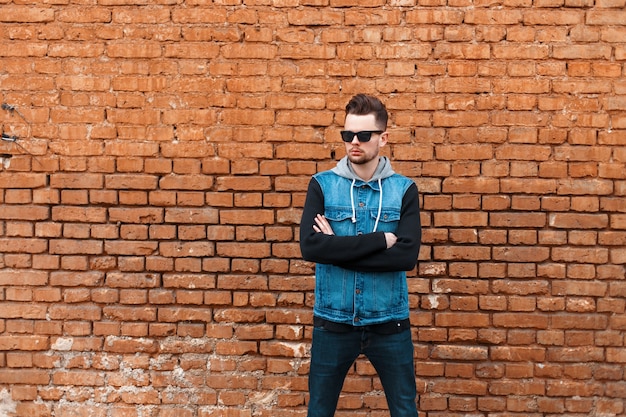 Hombre de moda hipster en ropa de jeans con gafas de sol cerca de una pared de ladrillos