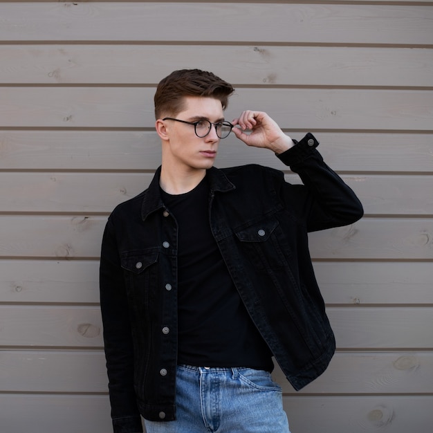 Hombre de moda joven endereza gafas con estilo cerca de la pared de madera en la calle