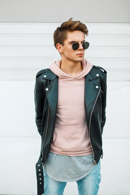 Foto hombre de moda joven guapo con un corte de pelo en gafas de sol en una chaqueta de cuero negro y una sudadera rosa cerca de una pared de metal blanco