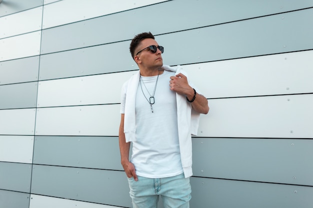 Hombre de moda joven inconformista con peinado de moda en gafas de sol en jeans en una camiseta blanca posando cerca de edificio moderno