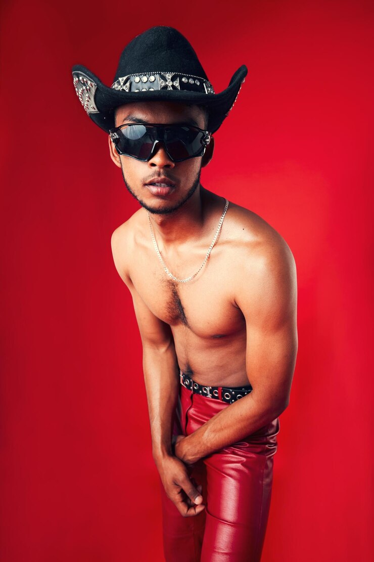 Hombre de moda y sombrero de vaquero gafas de sol y ropa sin camisa y gen z aislado sobre fondo rojo Modelo seguro pantalones frescos y de cuero con accesorios de gafas estética occidental en un estudio
