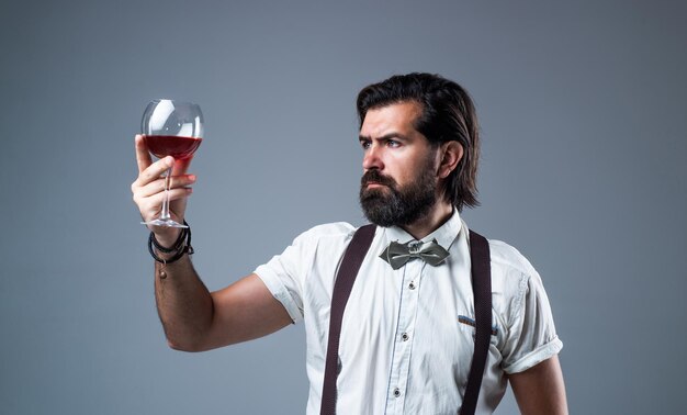 Hombre de moda en tirantes y pajarita con vino seguro de hombre elegante bebiendo vino de vidrio bebida alcohólica elegante sommelier degustación de bebidas enólogo hipster barbudo a la antigua