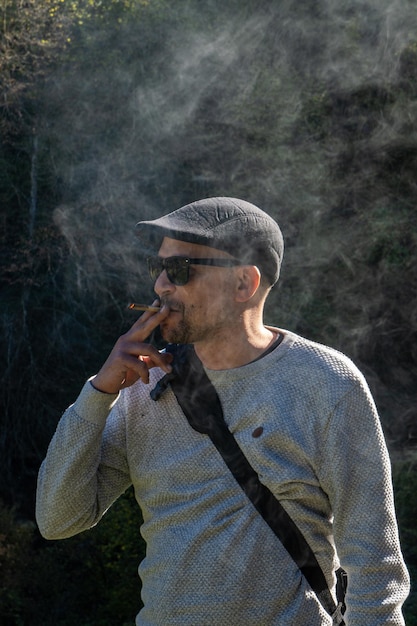 Hombre moderno y artista con una gorra bohemia con gafas de sol fumando su tabaco de liar iluminado