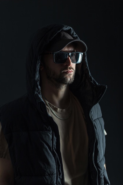 Un hombre moderno y elegante con una gorra y gafas de sol con ropa de moda en la oscuridad sobre un fondo negro