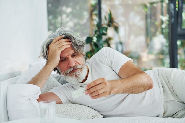 Hombre moderno con estilo senior enfermo con cabello gris y barba en el interior acostado en la cama y se siente mal