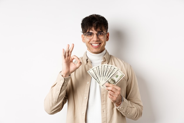 Hombre moderno guapo con gafas mostrando billetes de un dólar y gesto bien, haciendo dinero, de pie con efectivo en la pared blanca.