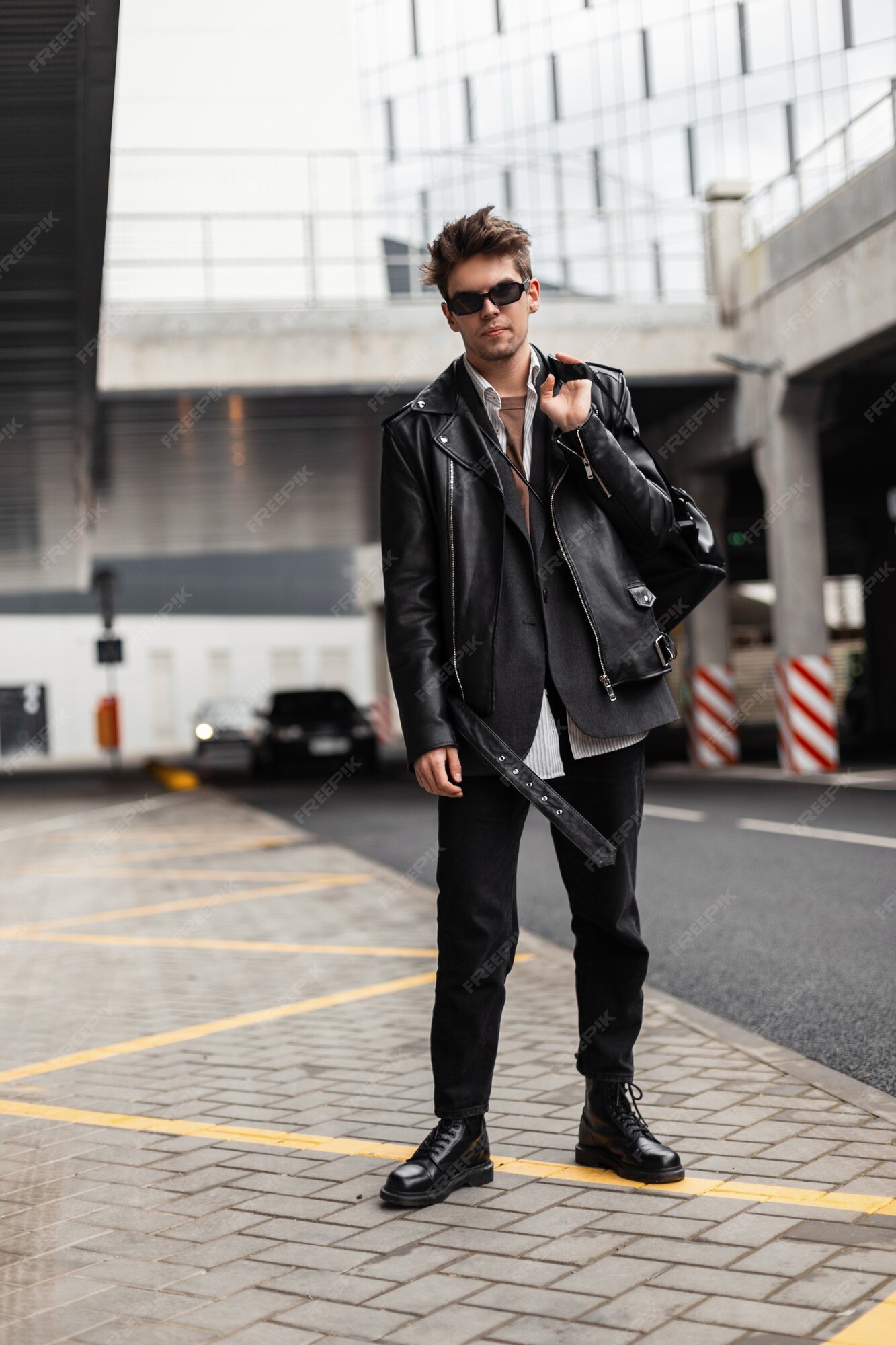 El hombre de moda inconformista ropa negra juvenil de moda en gafas de sol vintage con una elegante mochila se encuentra en la calle en un día primavera.