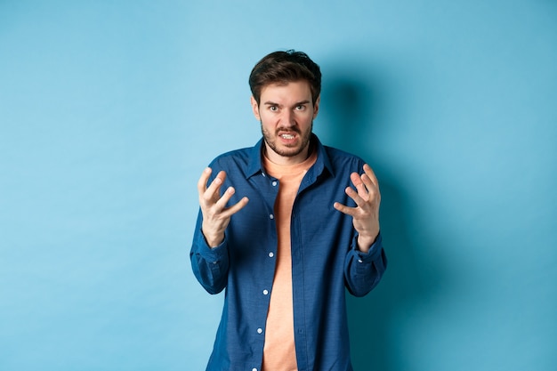 Foto hombre molesto y enojado que levanta las manos y aprieta los dientes indignados, mira cabreado a la cámara, va a matar a alguien, de pie sobre un fondo azul.