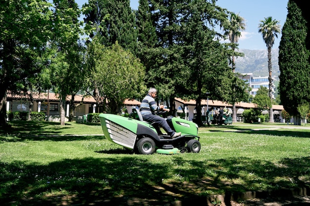 El hombre monta una cortadora de césped grande verde en el parque entre los árboles