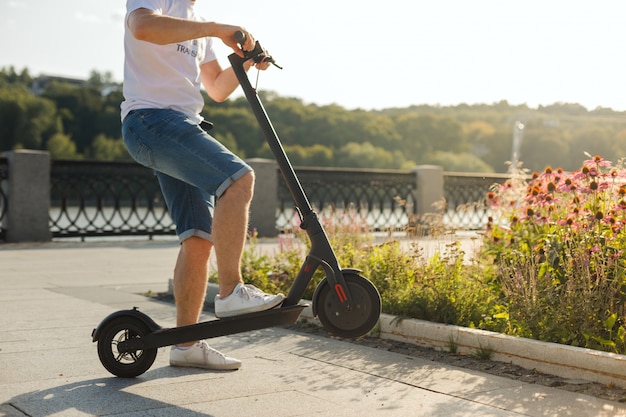 Hombre montando un scooter ecológico patada eléctrica en un parque en un clima soleado en las aceras. Luz tenue