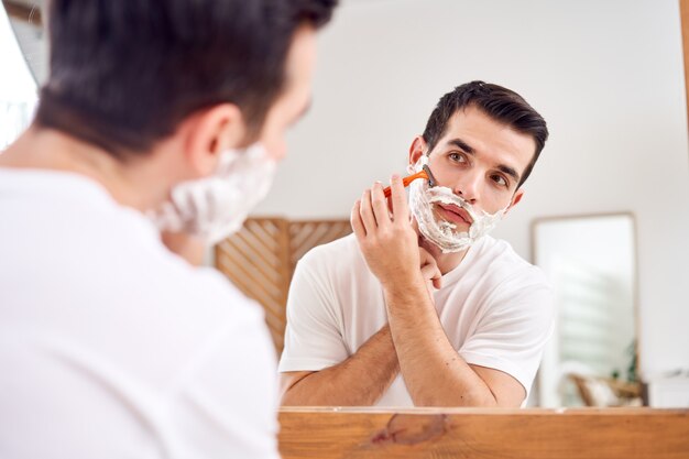 Hombre moreno en camiseta blanca se afeita mientras está de pie cerca del espejo en el baño en la mañana