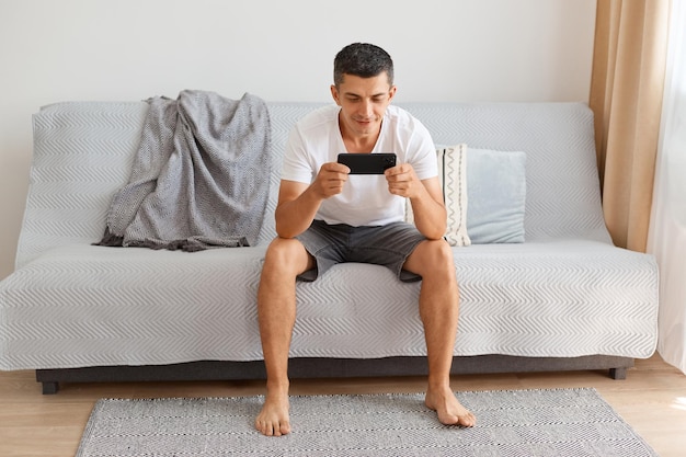 Hombre moreno caucásico concentrado vestido con camiseta blanca y pantalones cortos, posando en la sala de luz mientras está sentado en el sofá gris, jugando a través del teléfono celular, con expresión tranquila.