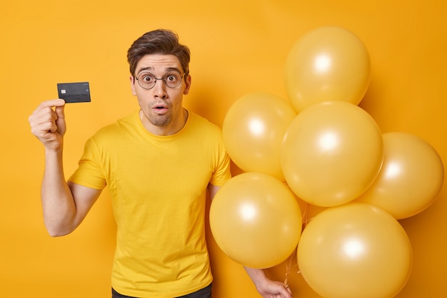 El hombre moreno impactado se siente impresionado vestido con una camiseta informal, tiene una tarjeta de crédito y un montón de globos inflados se prepara para un evento festivo aislado sobre fondo amarillo Concepto de ocasión festiva