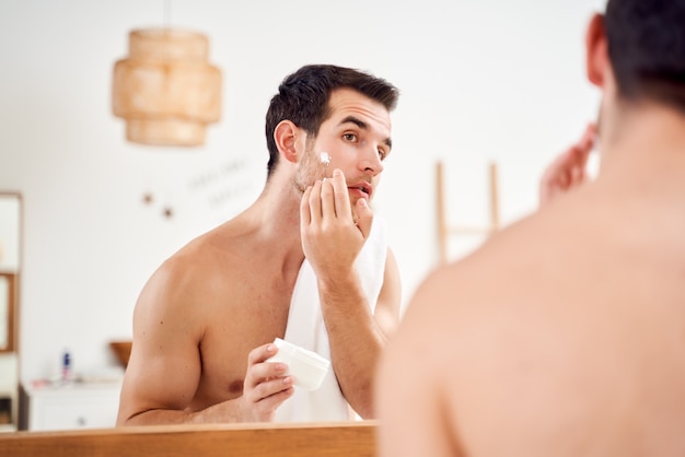 Foto hombre moreno con una toalla sobre los hombros con un tarro de crema en sus manos se encuentra frente al espejo del baño en la mañana