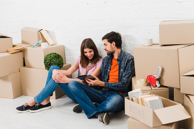 Foto hombre mostrando tableta digital a su esposa sentada entre las pilas de cajas de cartón