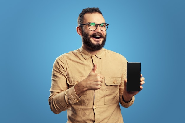 Hombre mostrando teléfono móvil con pantalla en blanco y mostrando el pulgar hacia arriba contra azul