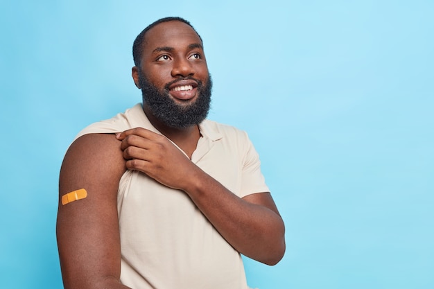 El hombre muestra el brazo después de recibir la vacuna promueve la campaña de vacunación pide mantenerse saludable y proteger su salud viste una camiseta informal en la pared azul