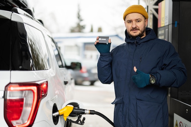 El hombre muestra la tarjeta de crédito mientras reabastece su automóvil SUV americano en la gasolinera cuando hace frío