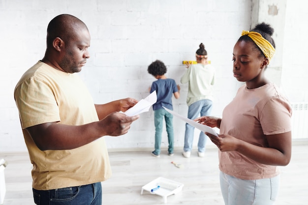 Hombre y mujer africana en ropa casual discutiendo papeles con instrucciones de montaje de muebles mientras sus dos hijos juegan junto a la pared