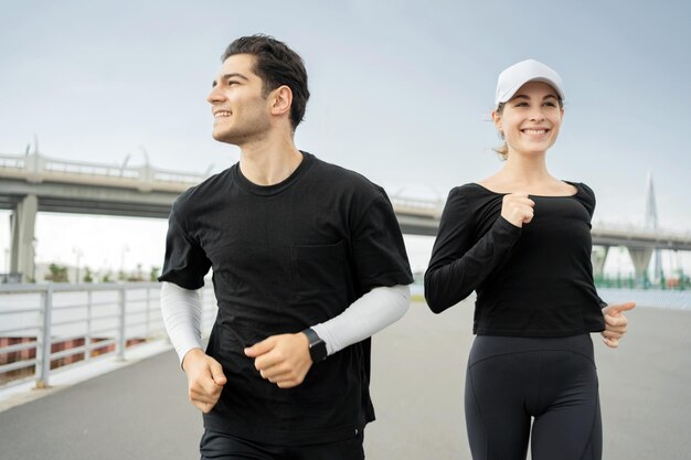 Hombre y mujer alegres corriendo juntos en un puente con ropa de fitness casual y relojes inteligentes