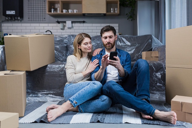 Un hombre y una mujer en un apartamento nuevo una pareja casada sentada en el suelo cerca de cajas de cartón usando el teléfono para comprar muebles en línea