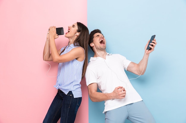 Hombre y mujer con auriculares cantando mientras escucha música en teléfonos inteligentes, aislado sobre pared colorida