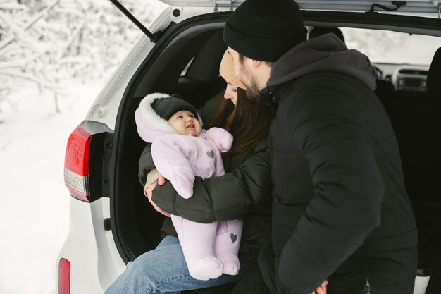 Un hombre, una mujer y un bebé están sentados en el maletero de un coche en un bosque cubierto de nieve en invierno