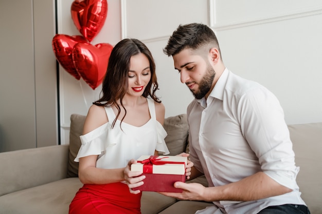 Hombre y mujer con caja de regalo y globos rojos en forma de corazón en el sofá en casa