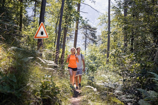 Hombre y mujer caminando en el bosque de garrapatas infectadas con señal de advertencia Riesgo de enfermedad de Lyme y transmitida por garrapatas