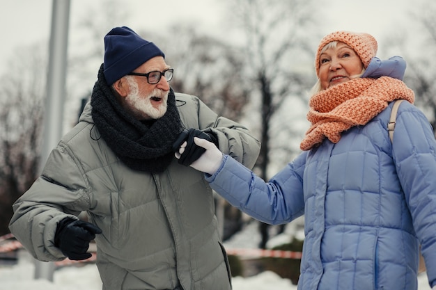 Hombre y mujer de edad positiva en ropa de invierno riendo y tomados de la mano mientras mantienen el equilibrio en la pista de patinaje