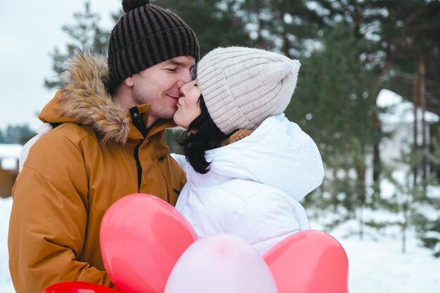 Un hombre y una mujer enamorados besándose en una cita al aire libre en invierno en la nieve con un regalo de globos rosas y rojos en forma de corazón. Día de San Valentín, amor, feliz pareja enamorada en un paseo.