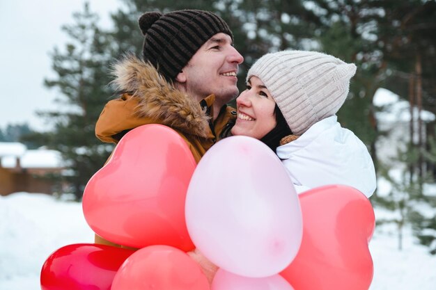 Un hombre y una mujer enamorados en una cita al aire libre en invierno en la nieve con un regalo de globos rosas y rojos en forma de corazón. Día de San Valentín, amor, feliz pareja enamorada en un paseo.