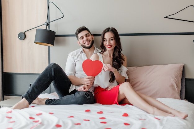 El hombre y la mujer enamorados de la tarjeta de San Valentín en forma de corazón rojo se sientan en la cama en su casa