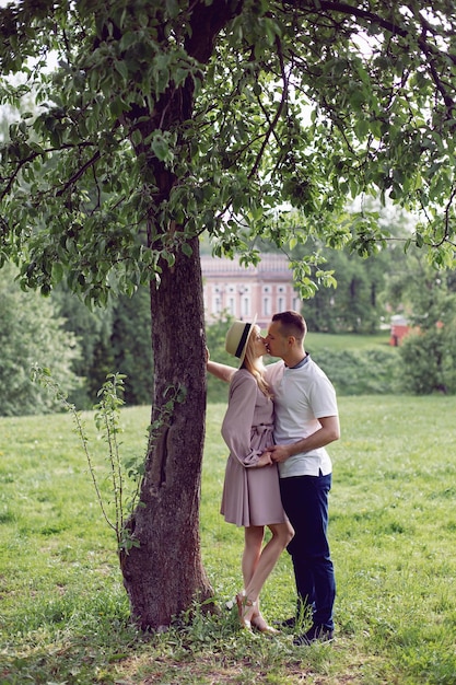 Un hombre y una mujer enamorados con un vestido y un sombrero están parados en un campo verde debajo de un árbol en verano