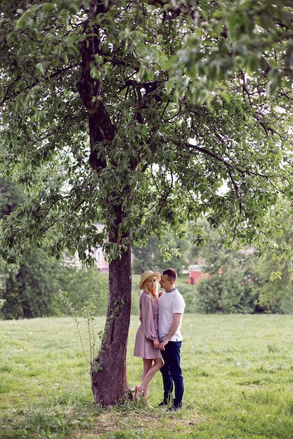 Un hombre y una mujer enamorados con un vestido y un sombrero están parados en un campo verde debajo de un árbol en verano