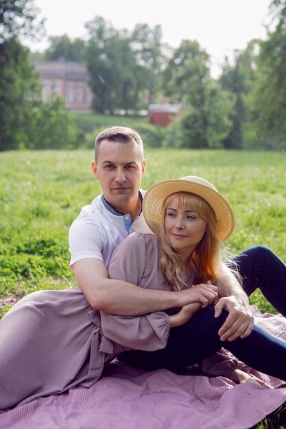 Un hombre y una mujer enamorados con un vestido y un sombrero están sentados en una manta en un campo verde debajo de un árbol en verano