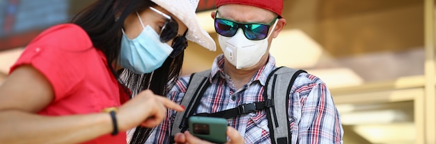 El hombre y la mujer en la estación de tren con máscaras médicas protectoras están buscando smartphon