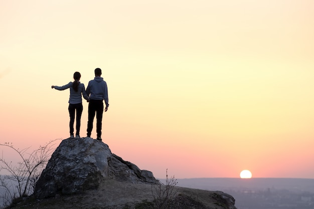 Hombre y mujer excursionistas de pie sobre la piedra grande al atardecer en las montañas. Pareja en roca alta en la naturaleza de noche. Concepto de turismo, viajes y estilo de vida saludable.