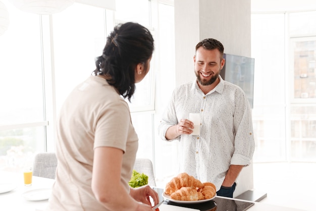 Hombre y mujer felices cocinando y desayunando en casa, mientras está de pie en la cocina luminosa
