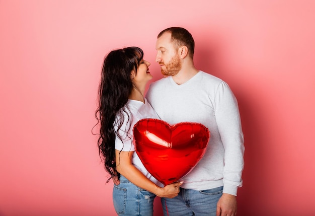 Un hombre y una mujer de fondo rosa sostienen en sus manos un gran globo inflable rojo en forma de corazón. Concepto de vacaciones - Día de San Valentín